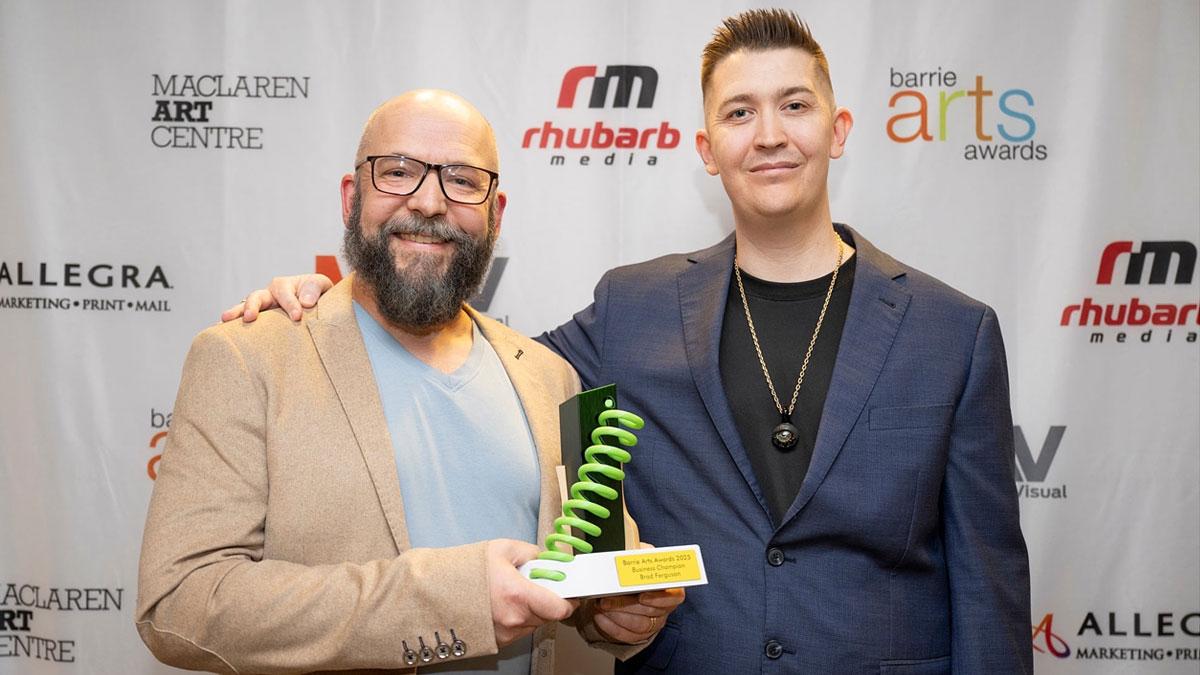 Two people standing in front of a backdrop with logos for Barrie Arts Awards, MacLaren Art Centre, Rhubarb Media, Allegra Barrie and MTAV. One of the people is holding an award.