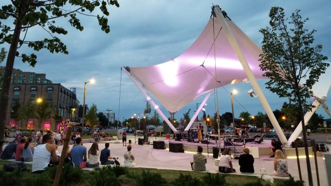 Outdoor venue covered bu roof. There is a crowd gathered around watching a performance.