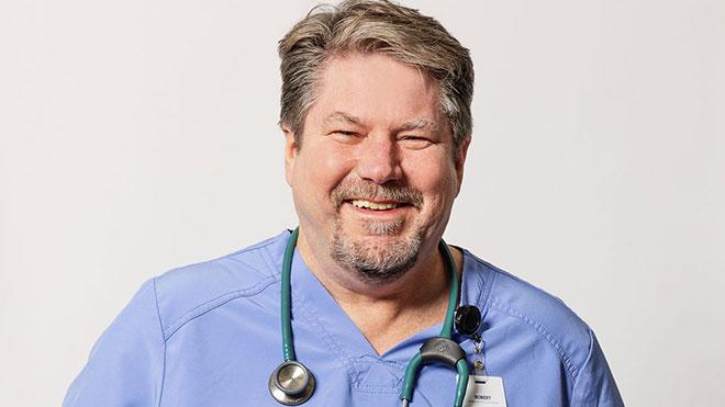 Robert dressed in scrubs, facing camera and smiling, grey background